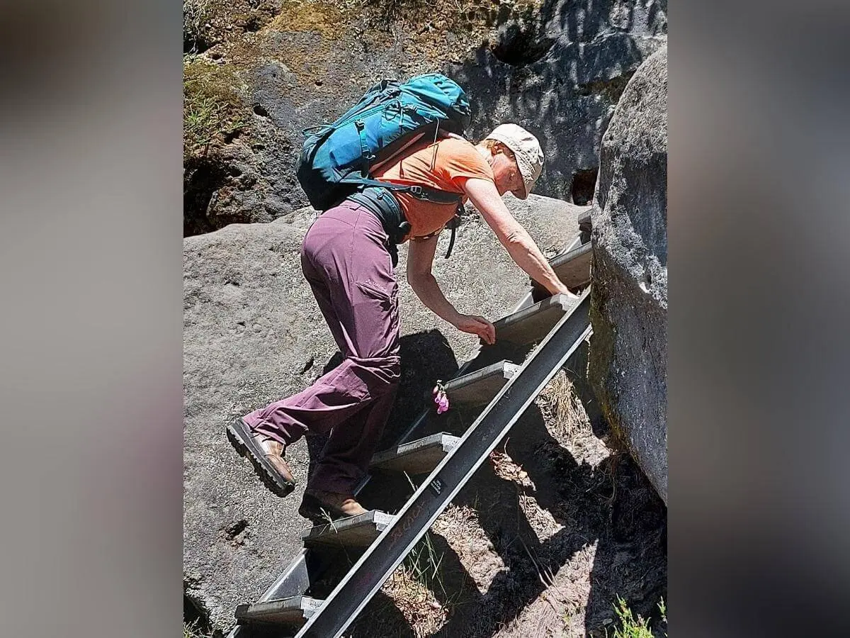 Person mit orangefarbenem Shirt und Rucksack erklimmt Metallleiter zwischen steilen Felsen. | © DAV Markt Schwaben | Foto Lutz Gründel