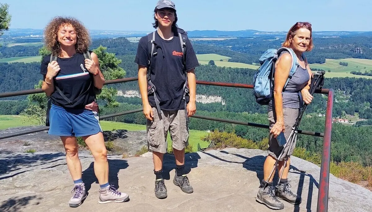 Drei Personen stehen auf Felsplateau mit Geländer, dahinter Tal mit Wald, Hügeln und blauem Himmel. | © DAV Markt Schwaben | Foto Lutz Gründel