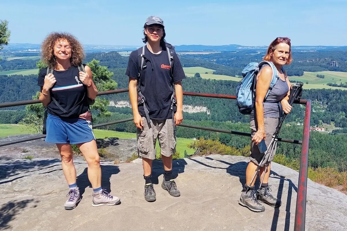 Drei Personen stehen auf Felsplateau mit Geländer, dahinter Tal mit Wald, Hügeln und blauem Himmel. | © DAV Markt Schwaben | Foto Lutz Gründel