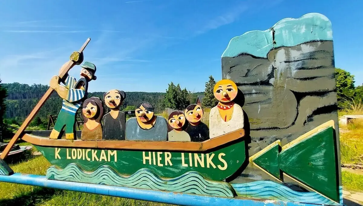 Buntes Holzschild zeigt Comicfiguren im Boot, Pfeil nach links, Felswand und Himmel im Hintergrund. | © DAV Markt Schwaben | Foto Angelika Göthe-Volkert