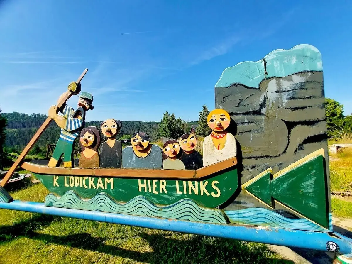 Buntes Holzschild zeigt Comicfiguren im Boot, Pfeil nach links, Felswand und Himmel im Hintergrund. | © DAV Markt Schwaben | Foto Angelika Göthe-Volkert