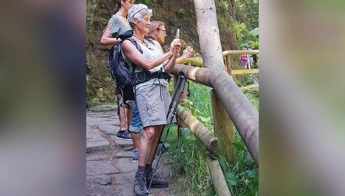 Drei Personen auf Waldpfad, vorne mit Smartphone und Wanderstöcken, umgeben von Bäumen und Sonnenlicht. | © DAV Markt Schwaben | Foto Lutz Gründel