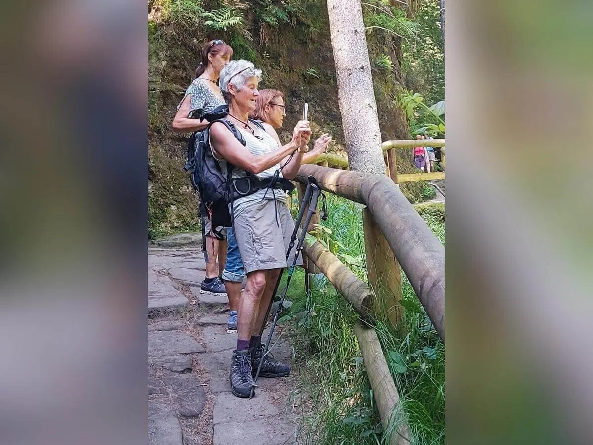 Drei Personen auf Waldpfad, vorne mit Smartphone und Wanderstöcken, umgeben von Bäumen und Sonnenlicht. | © DAV Markt Schwaben | Foto Lutz Gründel