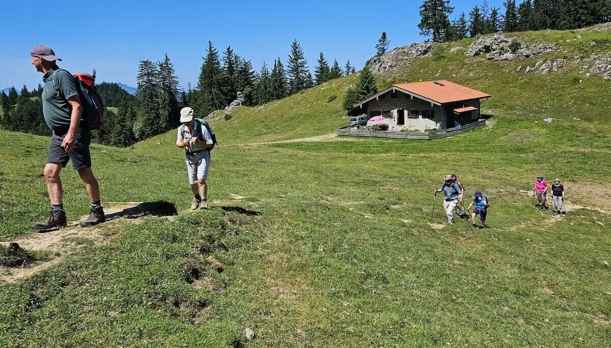 Fünf ältere Wanderer steigen bei sonnigem Wetter einen grasbewachsenen Hügel hinauf, Richtung Berghütte mit rotem Dach. | © DAV Markt Schwaben | Foto Hubert Inhofer