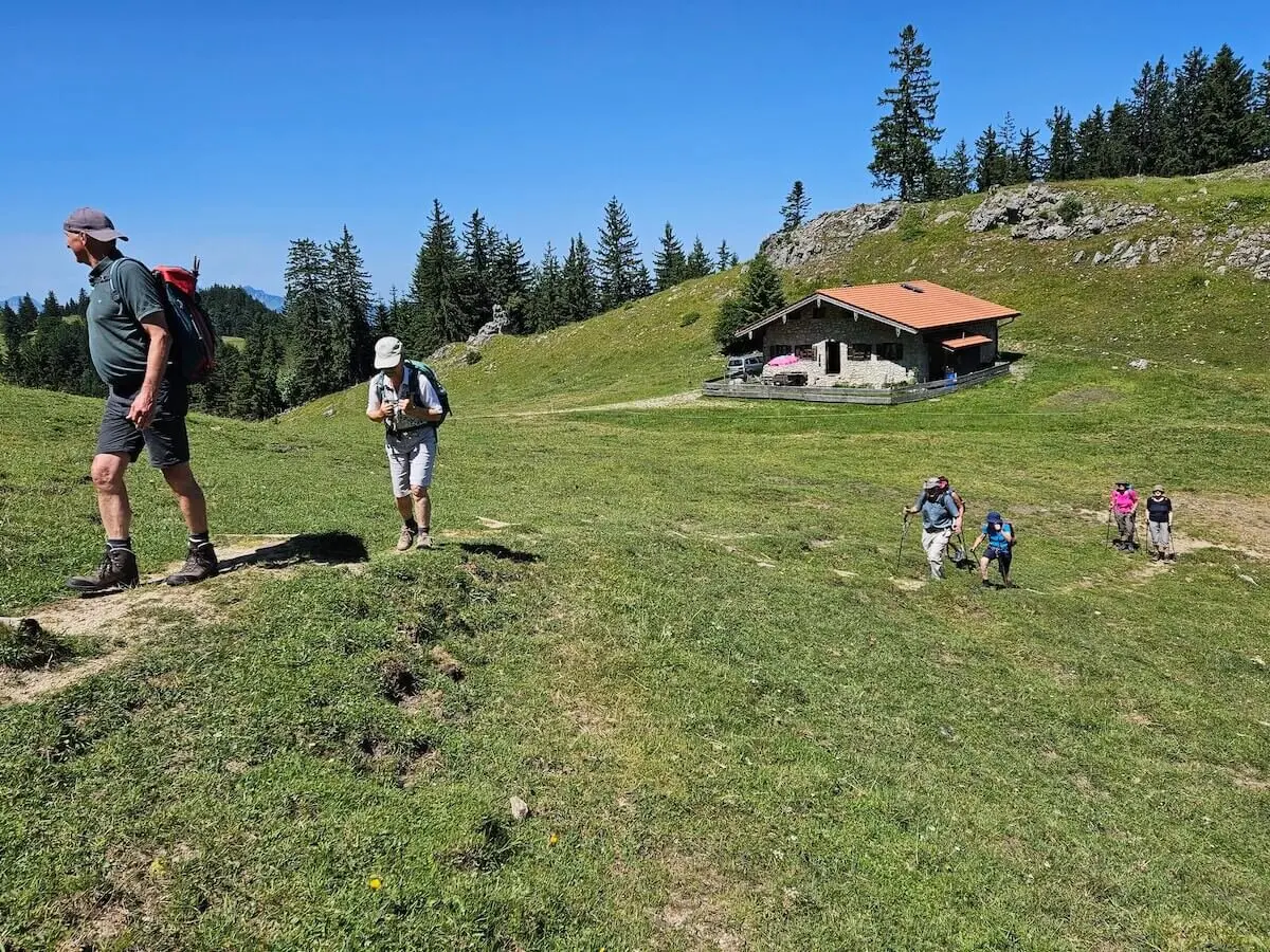 Fünf ältere Wanderer steigen bei sonnigem Wetter einen grasbewachsenen Hügel hinauf, Richtung Berghütte mit rotem Dach. | © DAV Markt Schwaben | Foto Hubert Inhofer