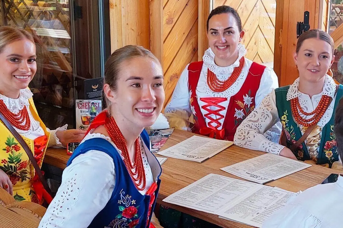 Vier Personen in bunter polnischer Festtracht sitzen an einem Holztisch in rustikalem Raum und blättern durch Speisekarten. | © DAV Markt Schwaben | Foto Lutz Gründel