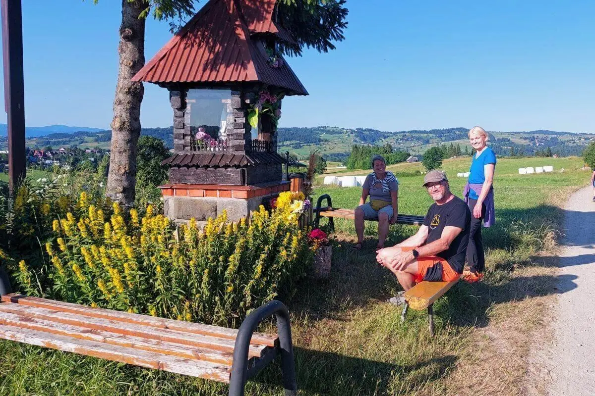 Drei Personen ruhen an einem blumengeschmückten Wegschrein mit rotem Dach; im Hintergrund Felder, Hügel und blauer Himmel. | © DAV Markt Schwaben | Foto Lutz Gründel