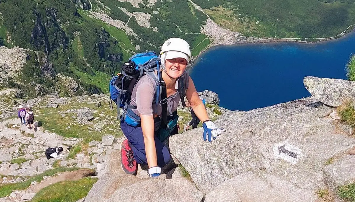 Eine Person mit Helm und Handschuhen steigt über felsigen Pfad; im Hintergrund ein Bergsee, grüne Hänge und weitere Wandernde. | © DAV Markt Schwaben | Foto Lutz Gründel