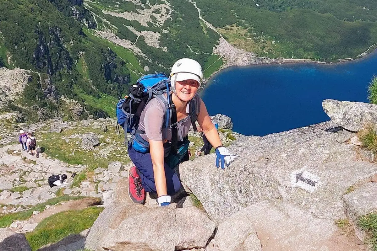 Eine Person mit Helm und Handschuhen steigt über felsigen Pfad; im Hintergrund ein Bergsee, grüne Hänge und weitere Wandernde. | © DAV Markt Schwaben | Foto Lutz Gründel