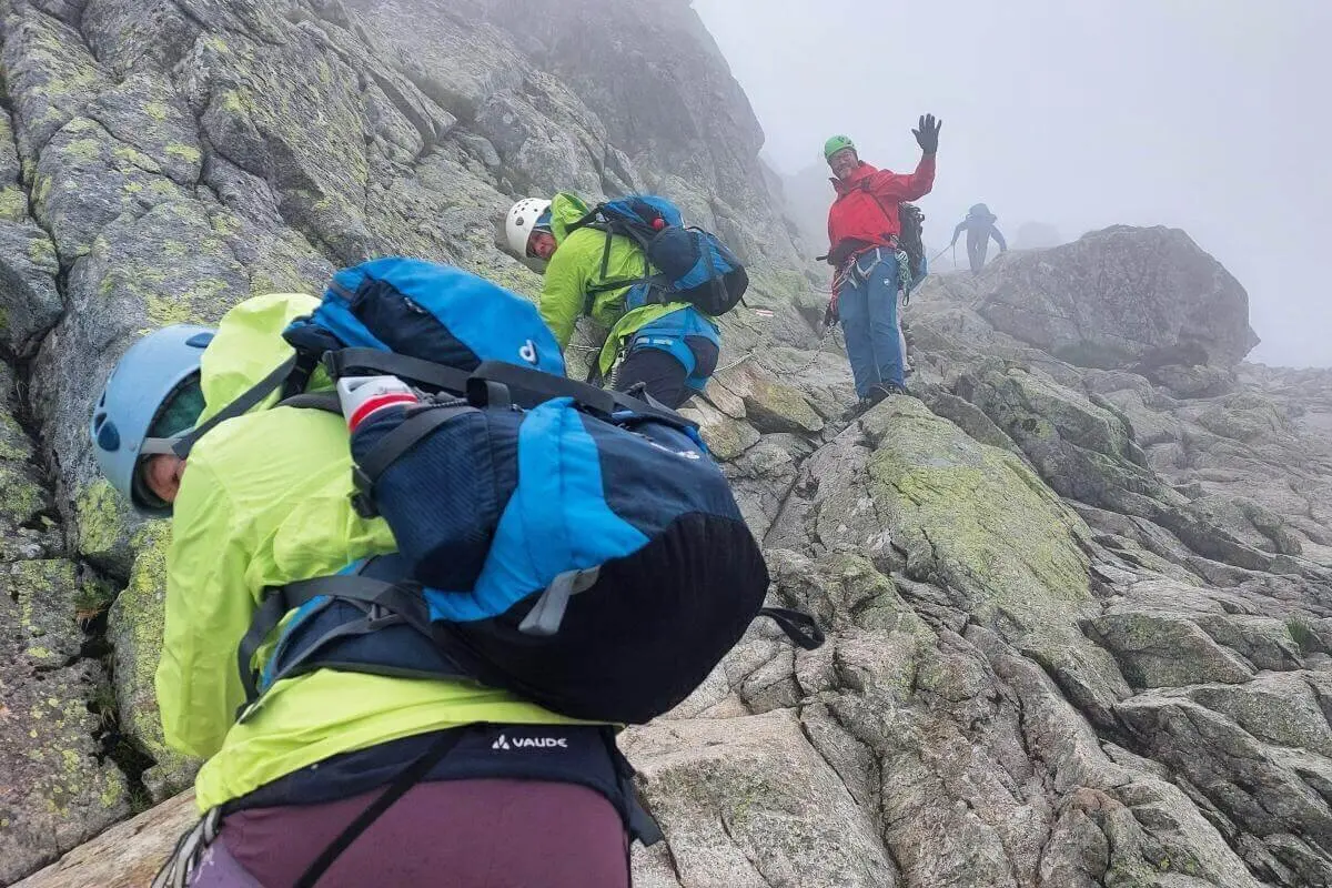 Mehrere Kletternde steigen mit Helm und Seil durch nebliges Felsgelände; eine Person winkt, andere sichern sich am steilen Hang. | © DAV Markt Schwaben | Foto Lutz Gründel