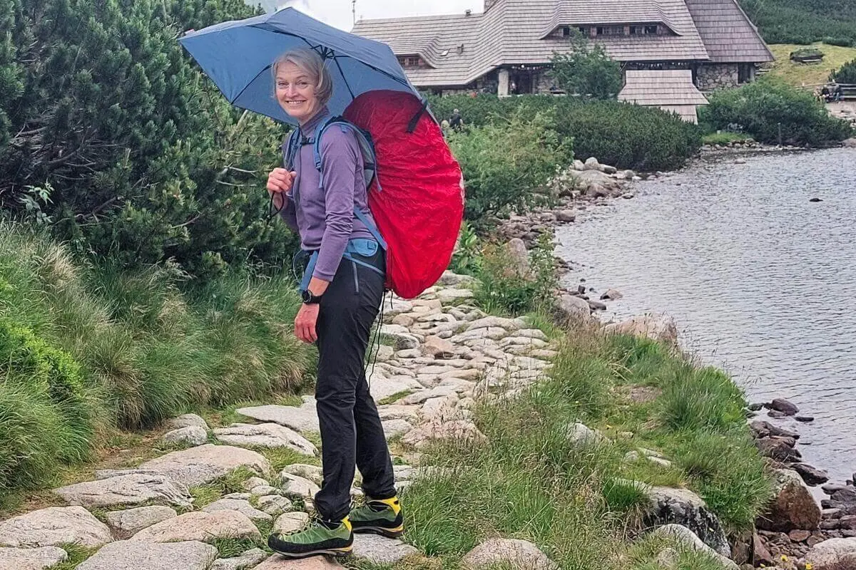 Eine Person mit rotem Rucksack und blauem Schirm wandert am See entlang; im Hintergrund ein Holzhaus. | © DAV Markt Schwaben | Foto Lutz Gründel