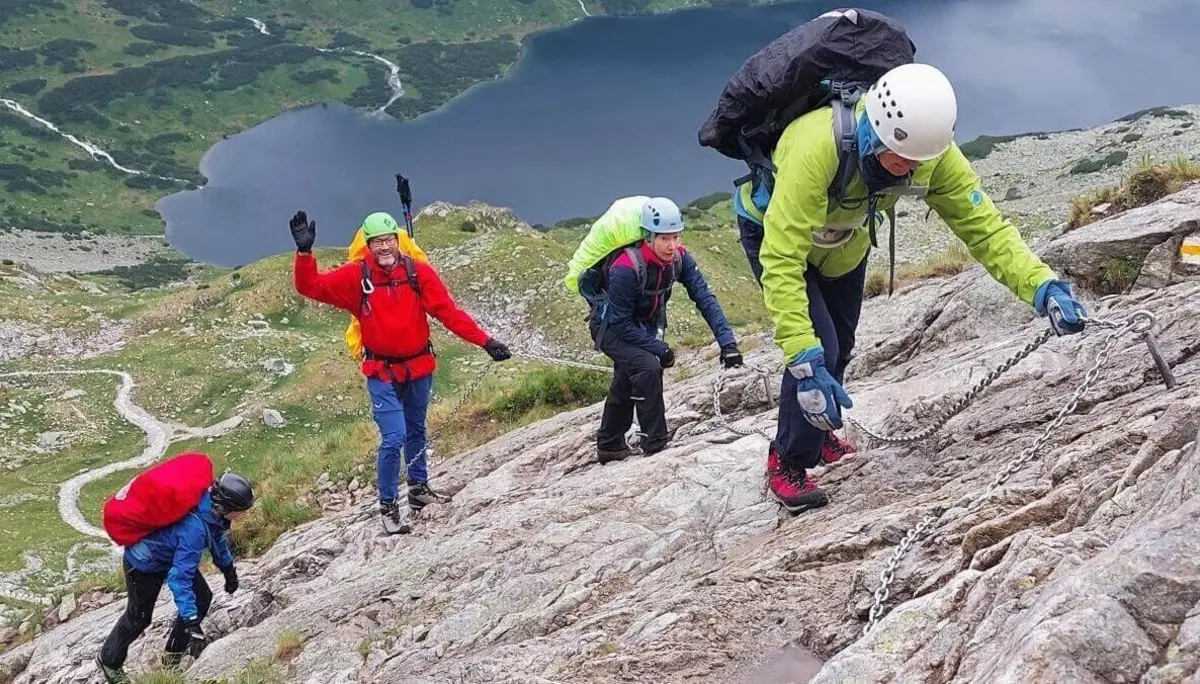 Fünf Kletternde steigen mit Helm und Kette über steilen Fels; im Hintergrund ein Bergsee und gewundene Pfade im Tal. | © DAV Markt Schwaben | Foto Lutz Gründel