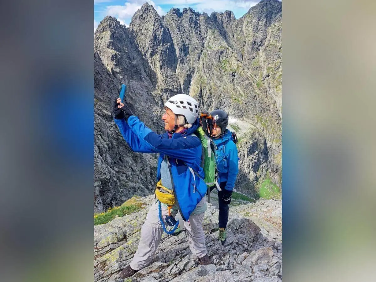 Zwei Kletternde mit Helm und Gurt stehen auf felsigem Berggrat; eine Person fotografiert, dahinter steile Gipfel und Wolken. | © DAV Markt Schwaben | Foto Lutz Gründel