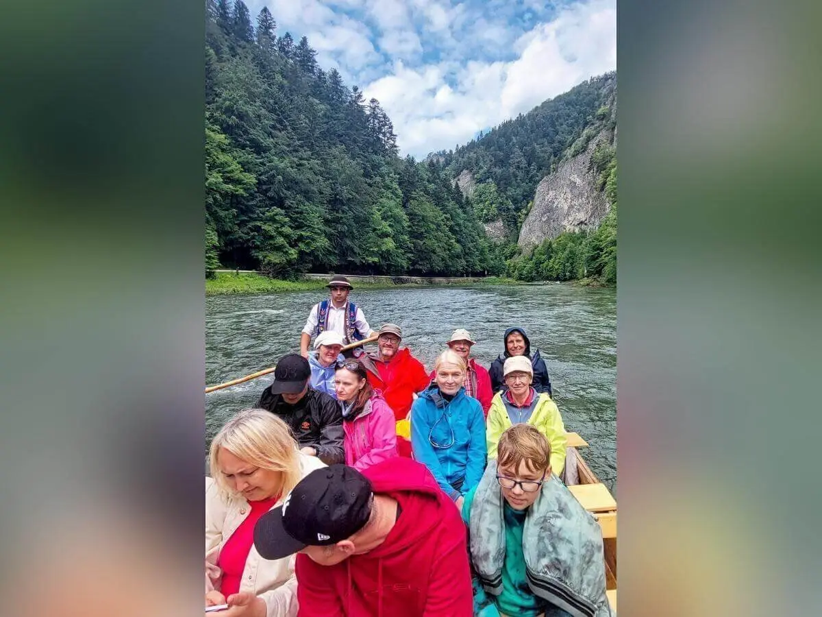 Eine Gruppe fährt in Regenjacken auf Holzboot durch ruhigen Fluss; hinten steht ein Guide in Tracht, umgeben von Wald und Felsen. | © DAV Markt Schwaben | Foto Lutz Gründel