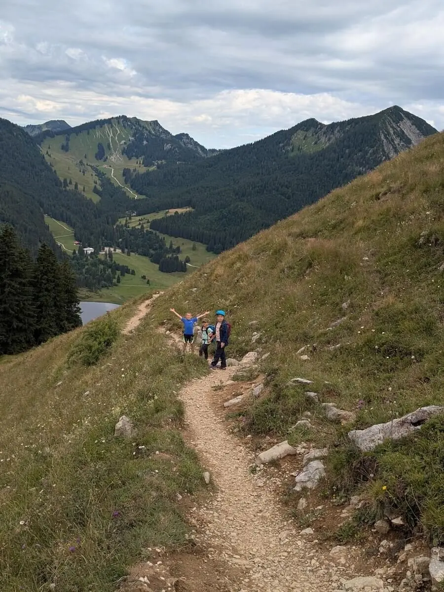 Drei Kinder wandern auf einem Bergpfad bei bewölktem Himmel. Sie gehen durch grünes Gelände mit Sicht auf umliegende Berge. | © DAV Markt Schwaben | Foto Familiengruppe