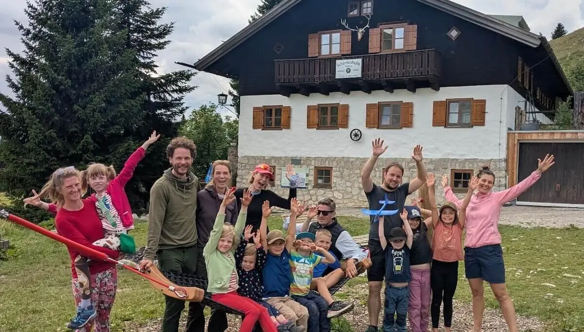 Fröhliche Familiengruppe posiert vor der Schönfeldhütte. Kinder und Erwachsene stehen lachend beisammen, einige winken. | © DAV Markt Schwaben | Foto Familiengruppe