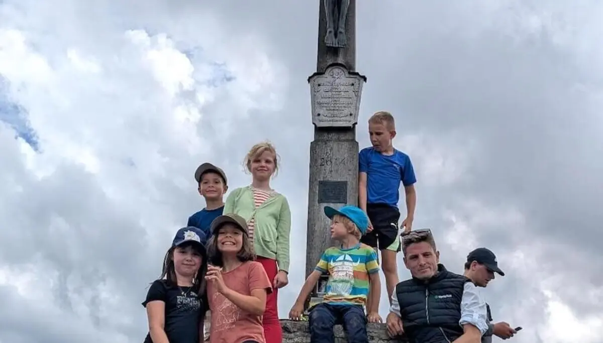 Familiengruppe steht am Gipfelkreuz des Jägerkamps mit Blick auf die umliegenden Berge. Kinder und Erwachsene lächeln in die Kamera. | © DAV Markt Schwaben | Foto Familiengruppe