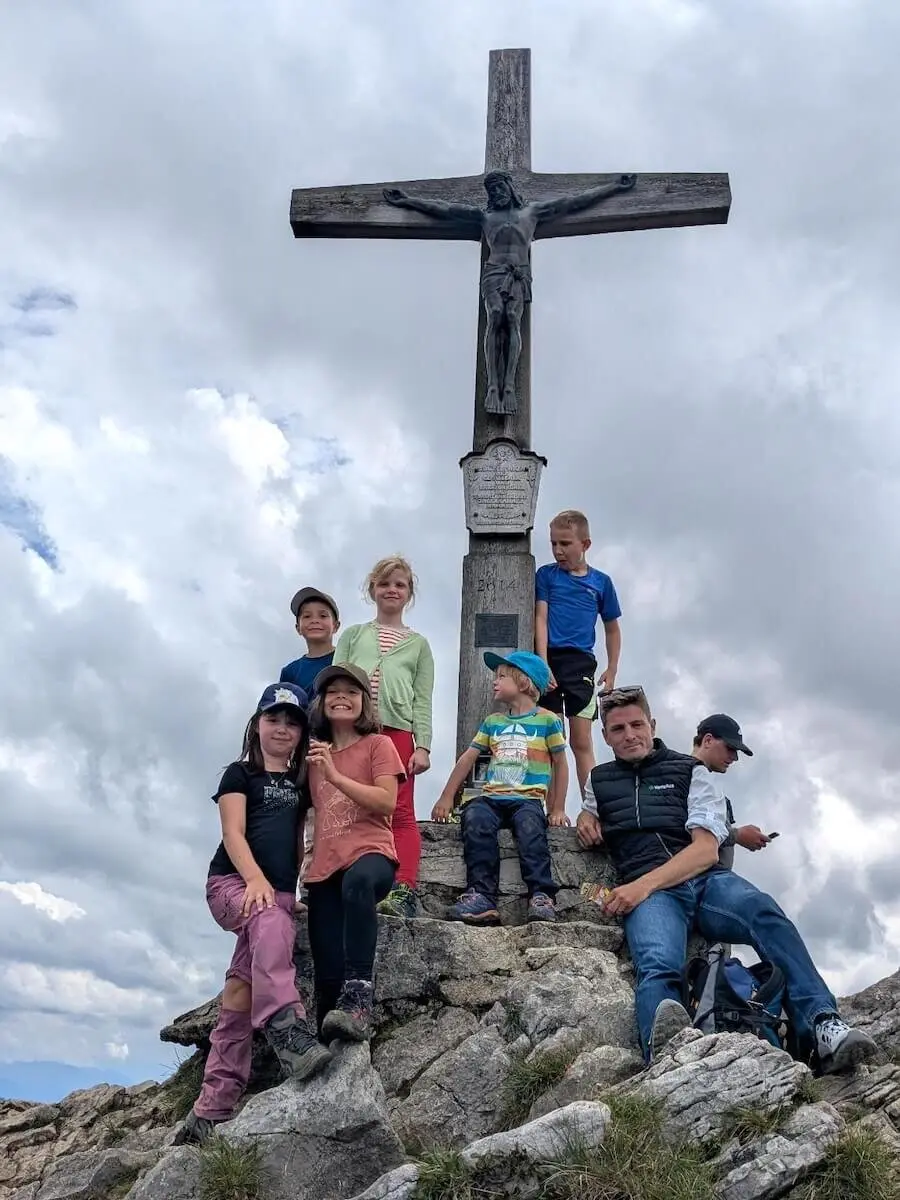 Familiengruppe steht am Gipfelkreuz des Jägerkamps mit Blick auf die umliegenden Berge. Kinder und Erwachsene lächeln in die Kamera. | © DAV Markt Schwaben | Foto Familiengruppe