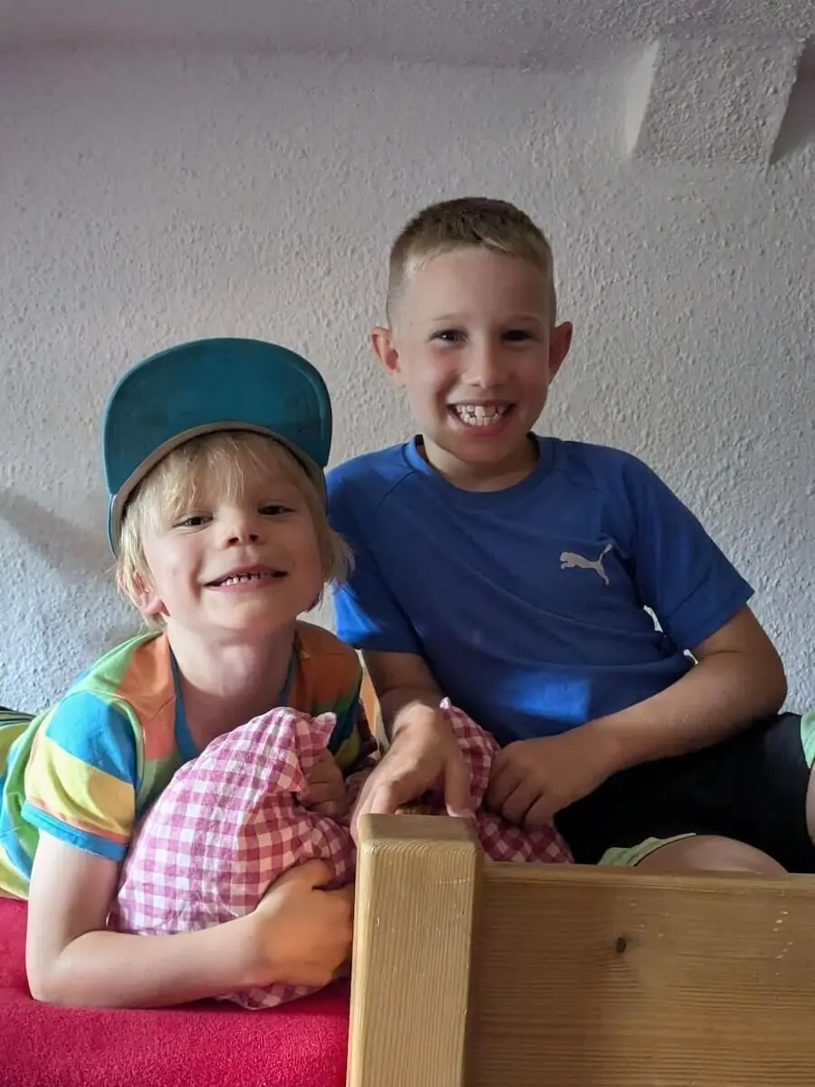 Zwei Kinder sitzen entspannt auf einem Holzbett mit roter Decke. Einer hält ein Kissen, beide genießen die Hüttenatmosphäre. | © DAV Markt Schwaben | Foto Familiengruppe