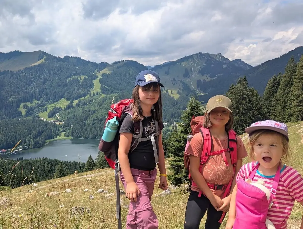 rei Kinder stehen auf einem grasbewachsenen Hang mit Rucksäcken. Im Hintergrund: Wälder, Berge und ein glitzernder See. | © DAV Markt Schwaben | Foto Familiengruppe