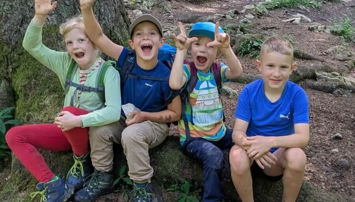 Vier Kinder sitzen auf einem freiliegenden Baumwurzelstock im Wald. Zwei winken, im Hintergrund wandern Erwachsene am Pfad entlang. | © DAV Markt Schwaben | Foto Familiengruppe