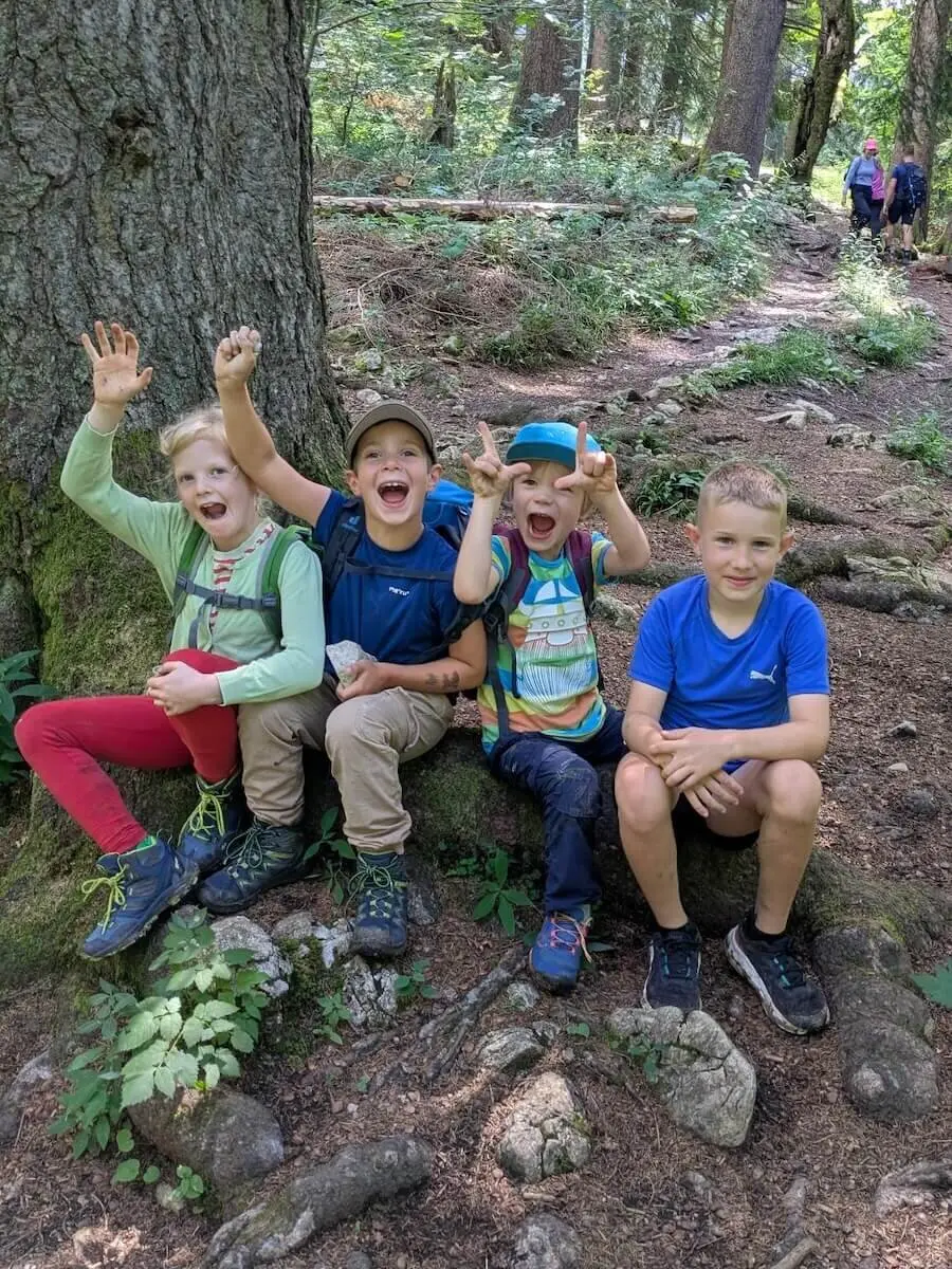 Vier Kinder sitzen auf einem freiliegenden Baumwurzelstock im Wald. Zwei winken, im Hintergrund wandern Erwachsene am Pfad entlang. | © DAV Markt Schwaben | Foto Familiengruppe