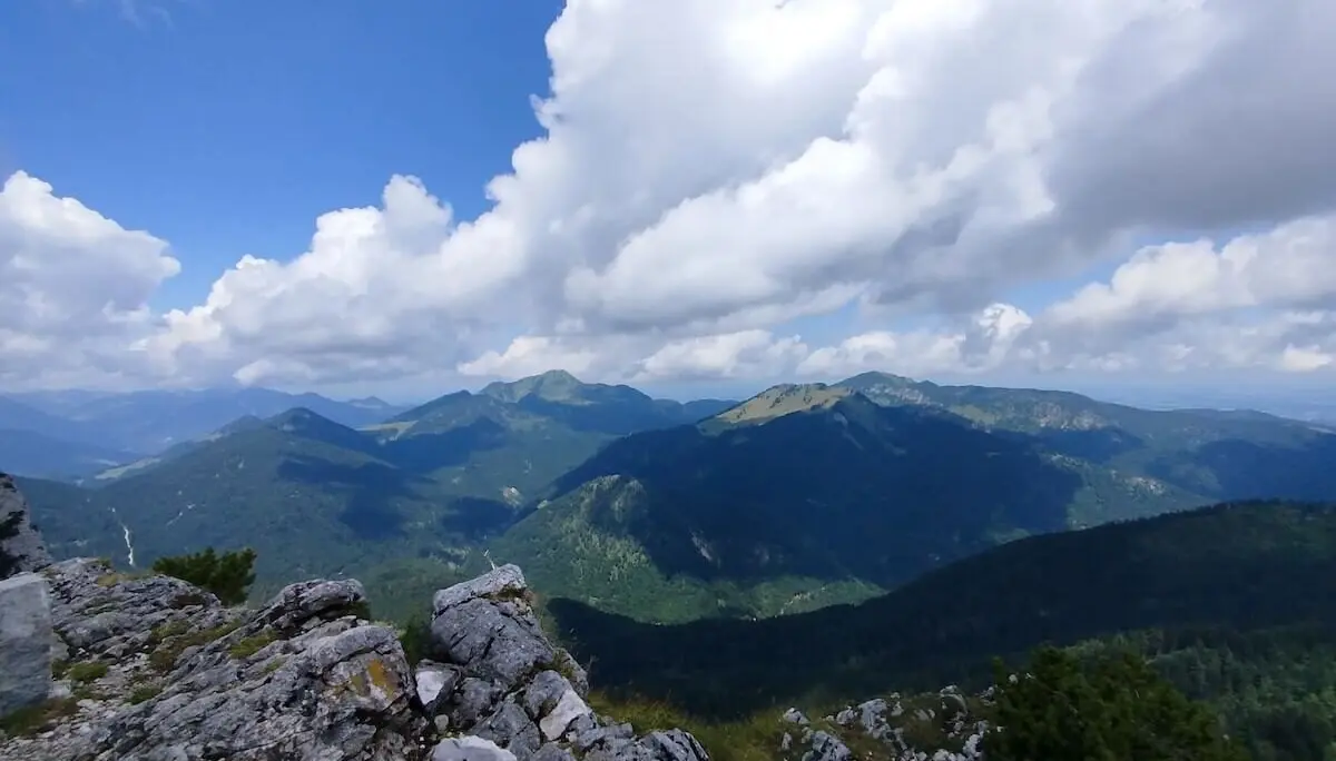 Felsen und Vegetation im Vordergrund, dahinter bewaldete Gipfel unter blauem Himmel mit weißen Wolken. | © DAV Markt Schwaben