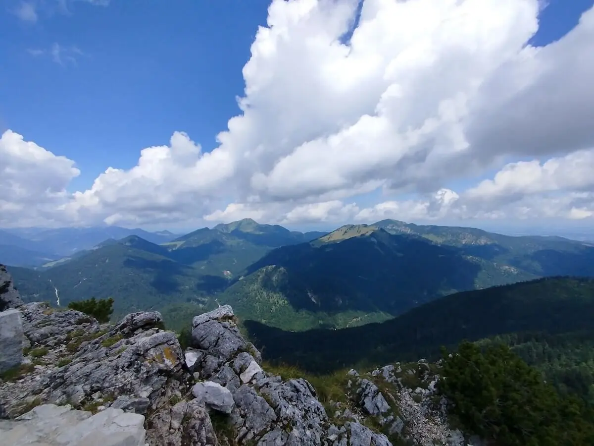 Felsen und Vegetation im Vordergrund, dahinter bewaldete Gipfel unter blauem Himmel mit weißen Wolken. | © DAV Markt Schwaben