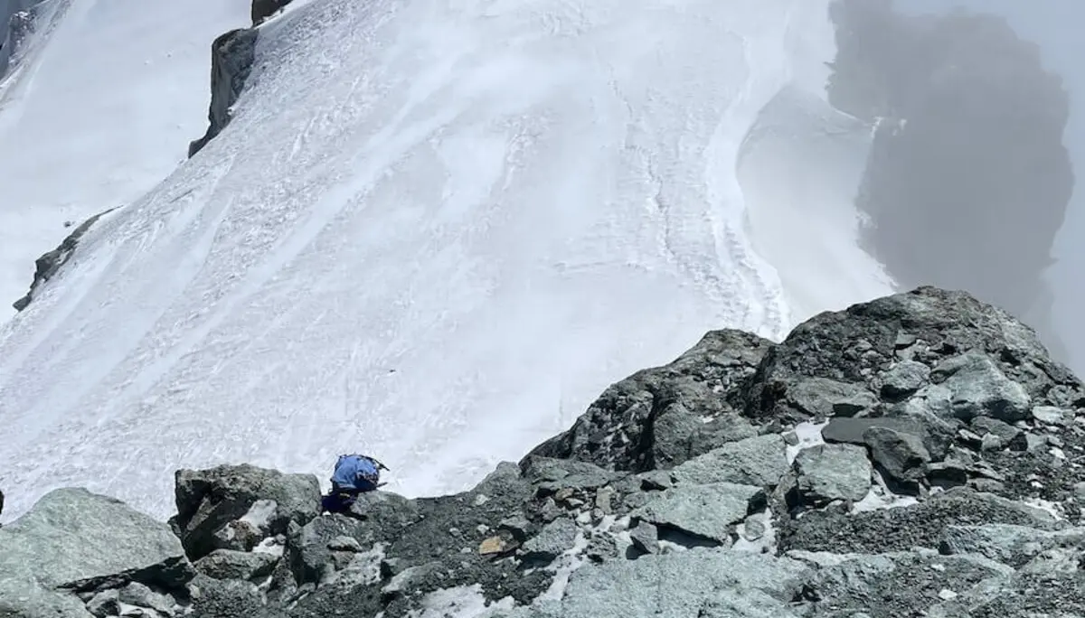 Ein Bergsteiger in blauer Jacke durchquert felsiges Gelände mit Schnee im Hochgebirge der Westalpen – Nebel am Gipfel. | © DAV Markt Schwaben | Christian Reischl