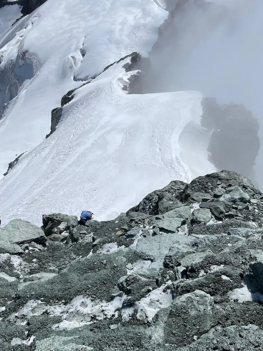 Ein Bergsteiger in blauer Jacke durchquert felsiges Gelände mit Schnee im Hochgebirge der Westalpen – Nebel am Gipfel. | © DAV Markt Schwaben | Christian Reischl