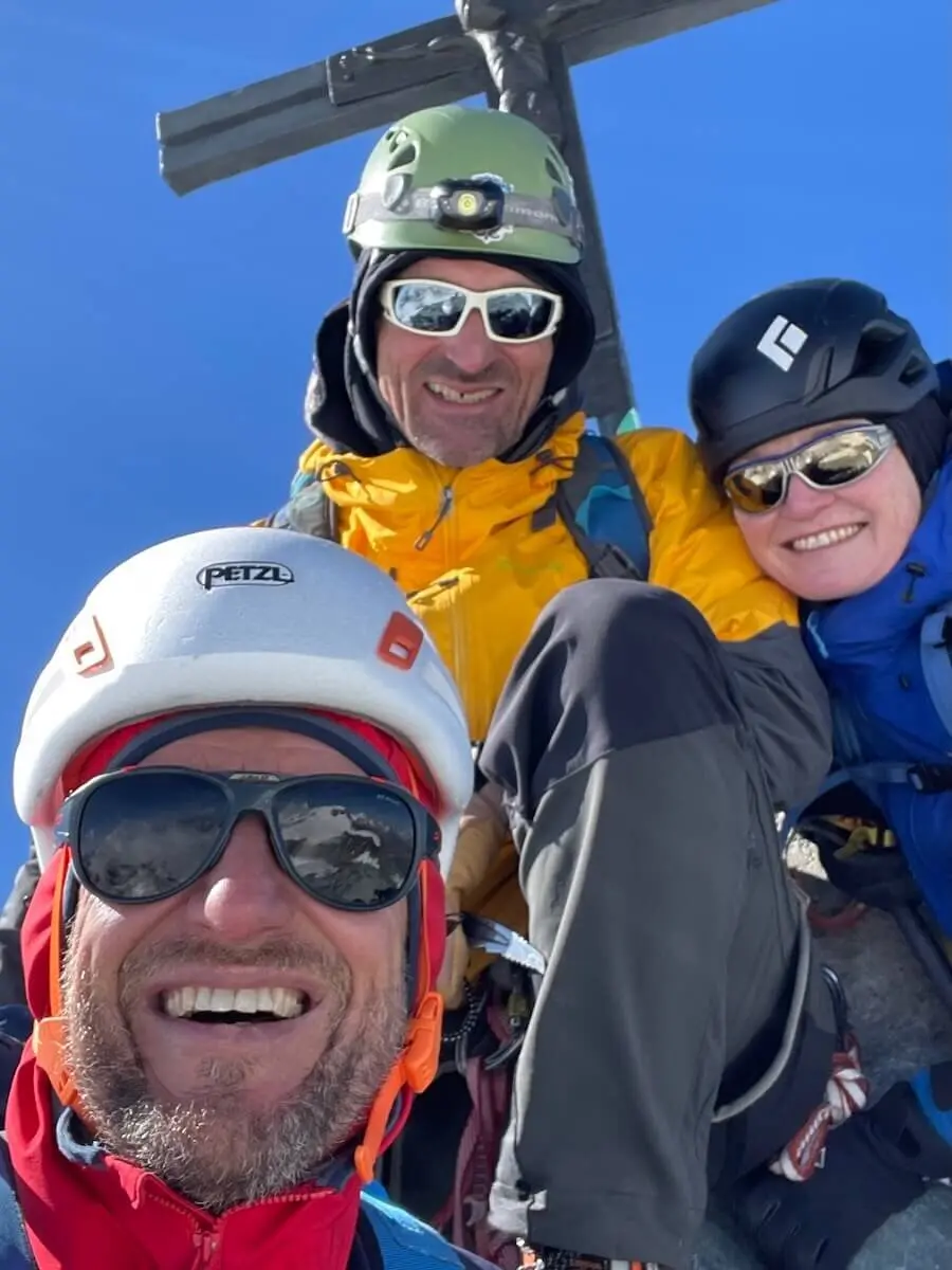 Drei Bergsteiger stehen vor einem Gipfelkreuz in den Westalpen – blauer Himmel und Kletterausrüstung unterstreichen das Ziel erreicht. | © DAV Markt Schwaben | Christian Reischl