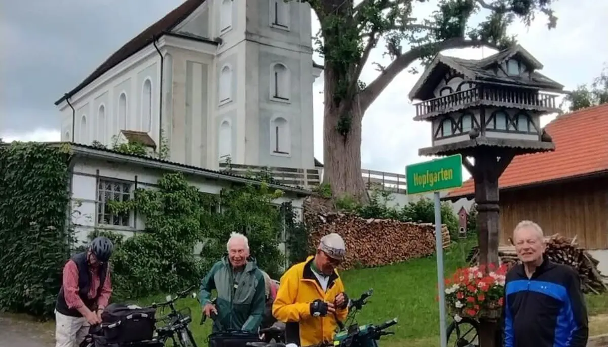 Fünf ältere Radfahrer stehen mit Gepäcktaschen vor einem Ortsschild – Helm, Kirche und Tourenausrüstung im Blick. | © DAV Markt Schwaben | Seniorengruppe