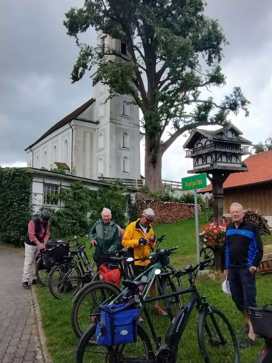 Fünf ältere Radfahrer stehen mit Gepäcktaschen vor einem Ortsschild – Helm, Kirche und Tourenausrüstung im Blick. | © DAV Markt Schwaben | Seniorengruppe