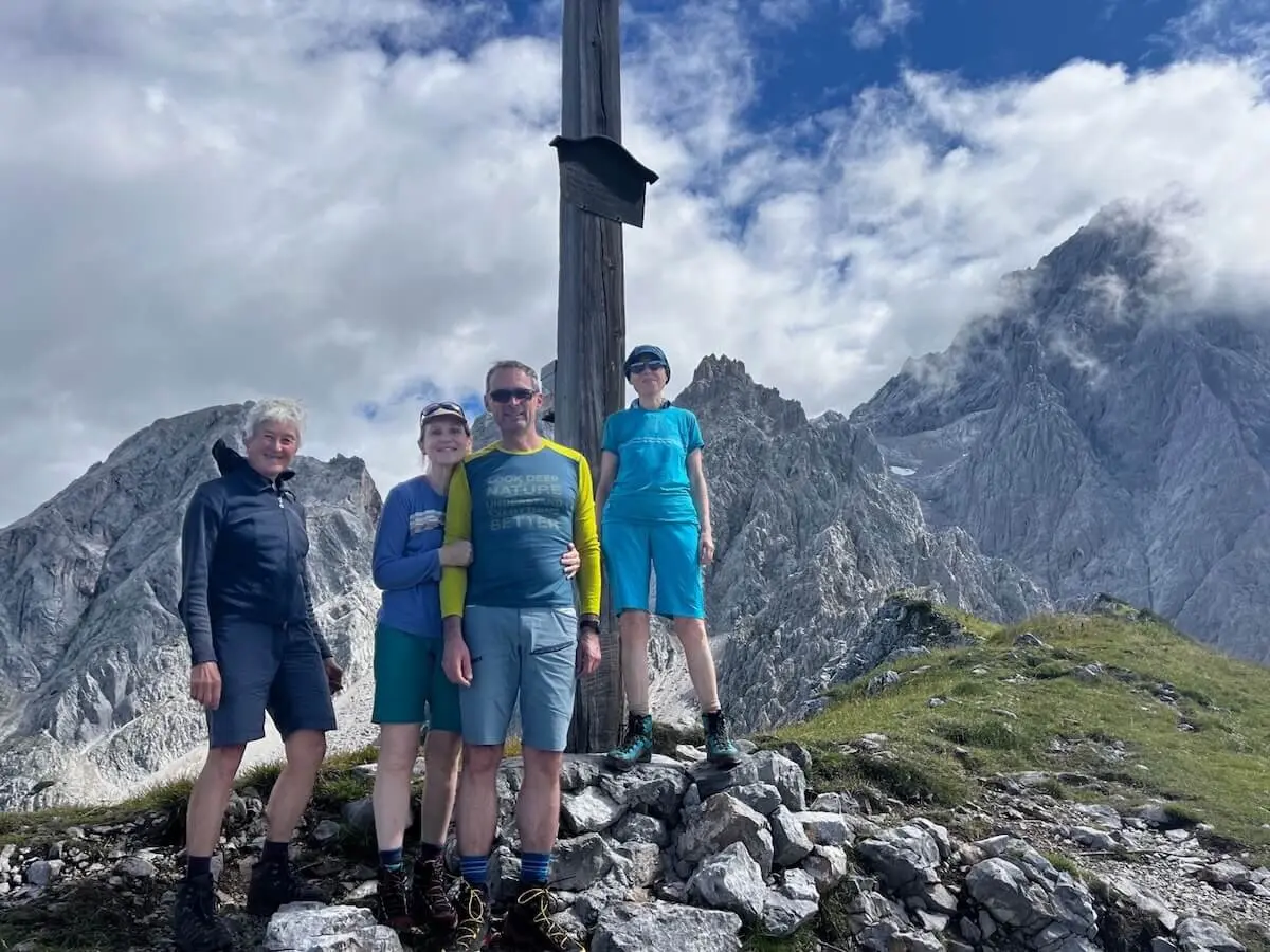 Vier Wandernde posieren vor einem Gipfelkreuz mit Blick auf felsige Bergspitzen unter teils bewölktem Himmel. | © DAV Markt Schwaben | Foto: Markus Sellmeier