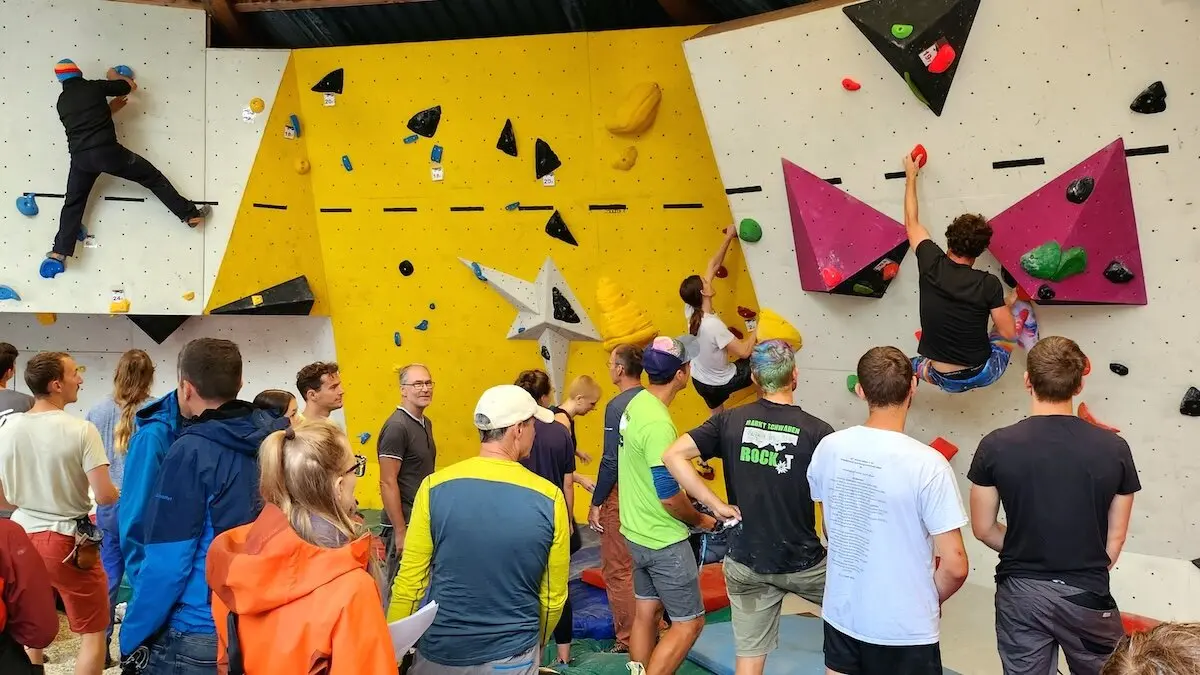 Mehrere Kletternde erklimmen die Boulderwand, während Zuschauer gespannt das Geschehen im Kletterzentrum verfolgen. | © DAV Markt Schwaben | Foto Anna Thoms