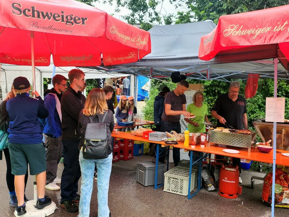 Besucher*innen stehen am Grillstand mit roten Schweiger-Schirmen, während Helfer Essen ausgeben – gesellige Stimmung im Freien. | © DAV Markt Schwaben | Foto Anna Thoms