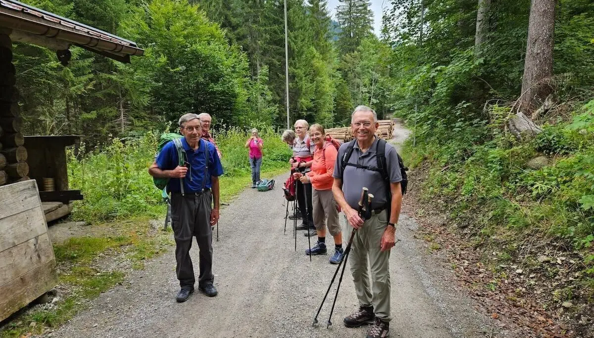 Sechs Teilnehmende der DAV-Wanderung stehen mit Stöcken auf einem Kiesweg im Wald, neben einer Holzhütte im Grünen. | © DAV Markt Schwaben | Foto Hubert Inhofer