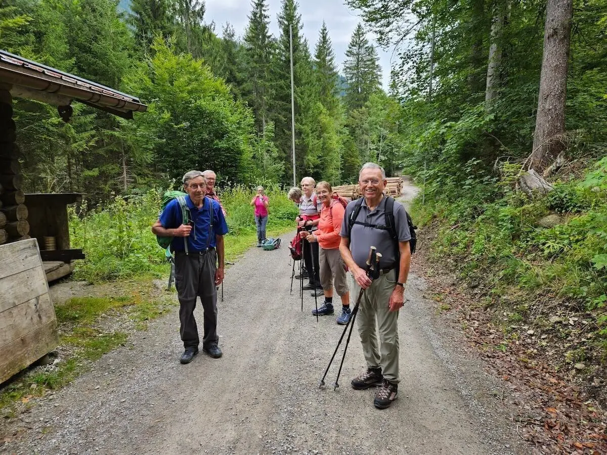 Sechs Teilnehmende der DAV-Wanderung stehen mit Stöcken auf einem Kiesweg im Wald, neben einer Holzhütte im Grünen. | © DAV Markt Schwaben | Foto Hubert Inhofer
