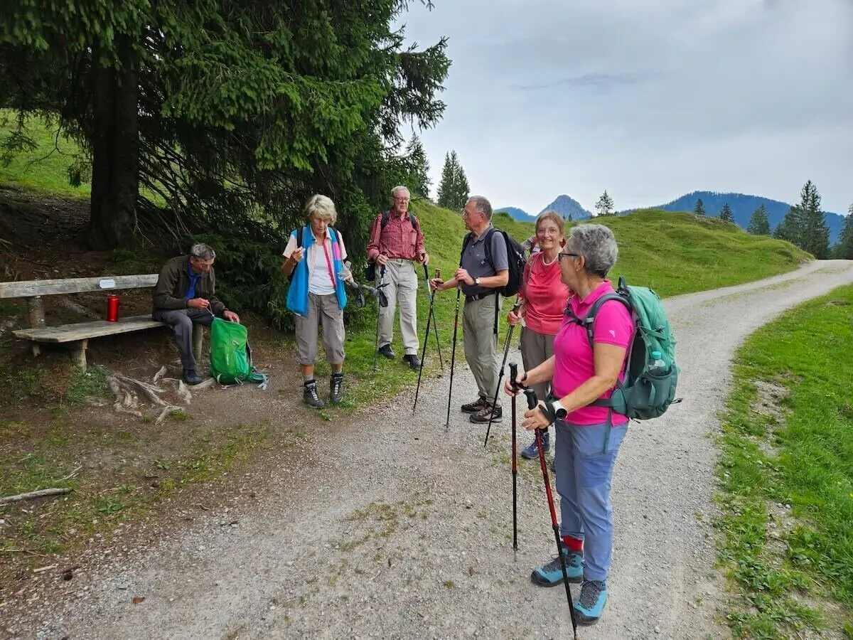 Fünf Wandernde stehen auf einem Bergpfad, eine Person sitzt auf einer Bank. Im Hintergrund: Wiesen, Wald und Bergpanorama. | © DAV Markt Schwaben | Foto Hubert Inhofer