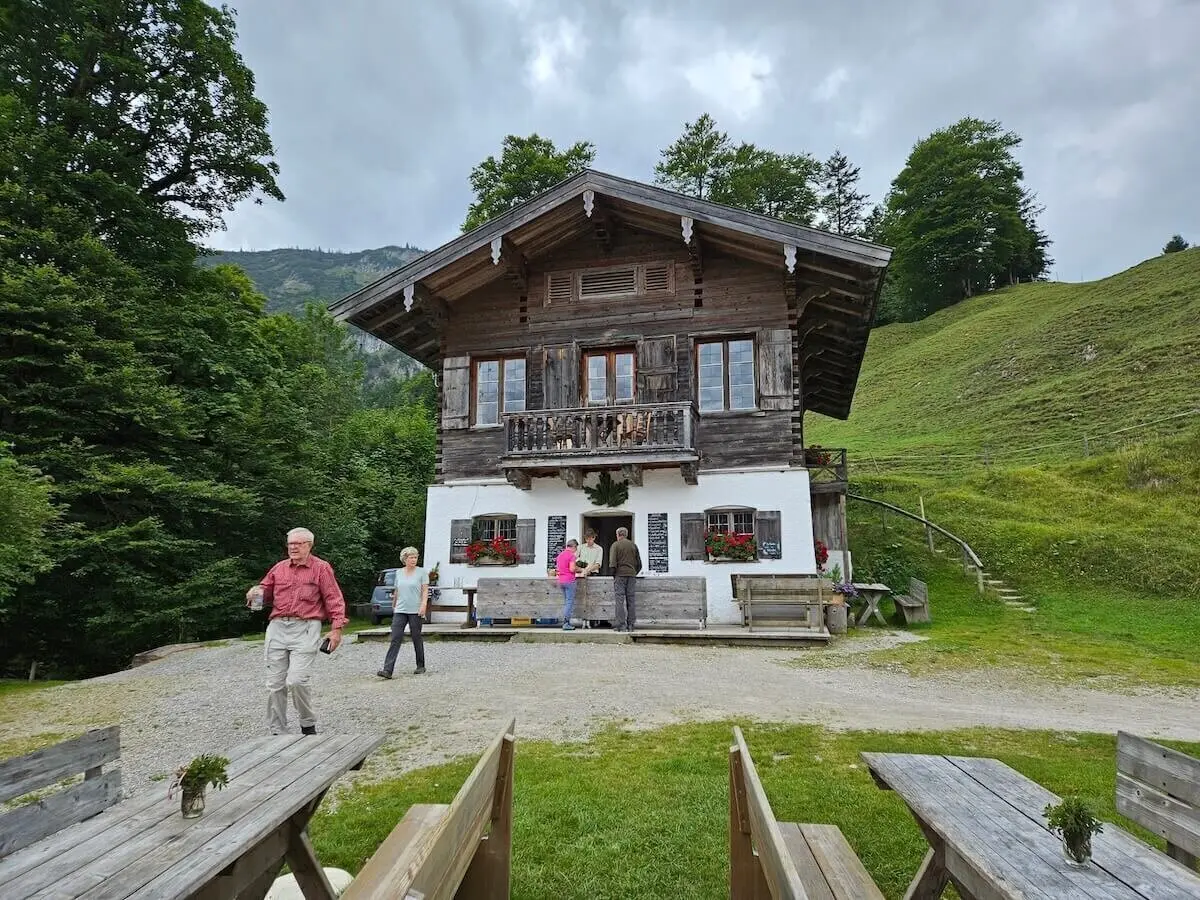 Mehrere Teilnehmende stehen vor der Königsalm mit Holzhütte, Blumenschmuck und Tischen – umgeben von alpiner Landschaft. | © DAV Markt Schwaben | Foto Hubert Inhofer