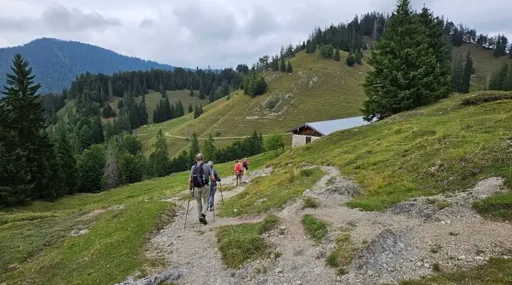 Drei Wandernde mit Rucksäcken und Stöcken auf einem Bergpfad, im Hintergrund eine Holzhütte und bewaldete Hänge. | © DAV Markt Schwaben | Foto Hubert Inhofer