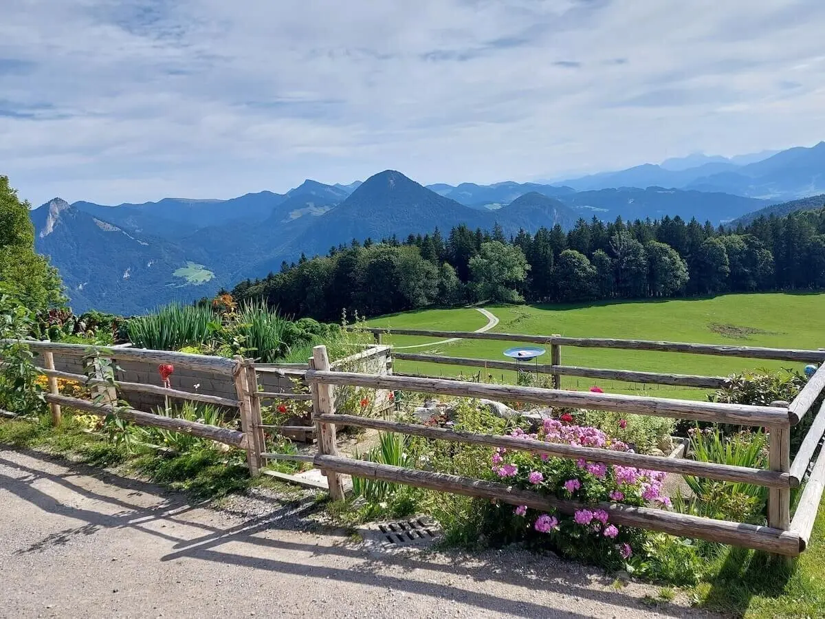 Im Vordergrund blühen bunte Blumen hinter einem Holzzaun, dahinter Wiese, Wald und Berggipfel unter Wolken. | © DAV Markt Schwaben | Foto Hubert Inhofer