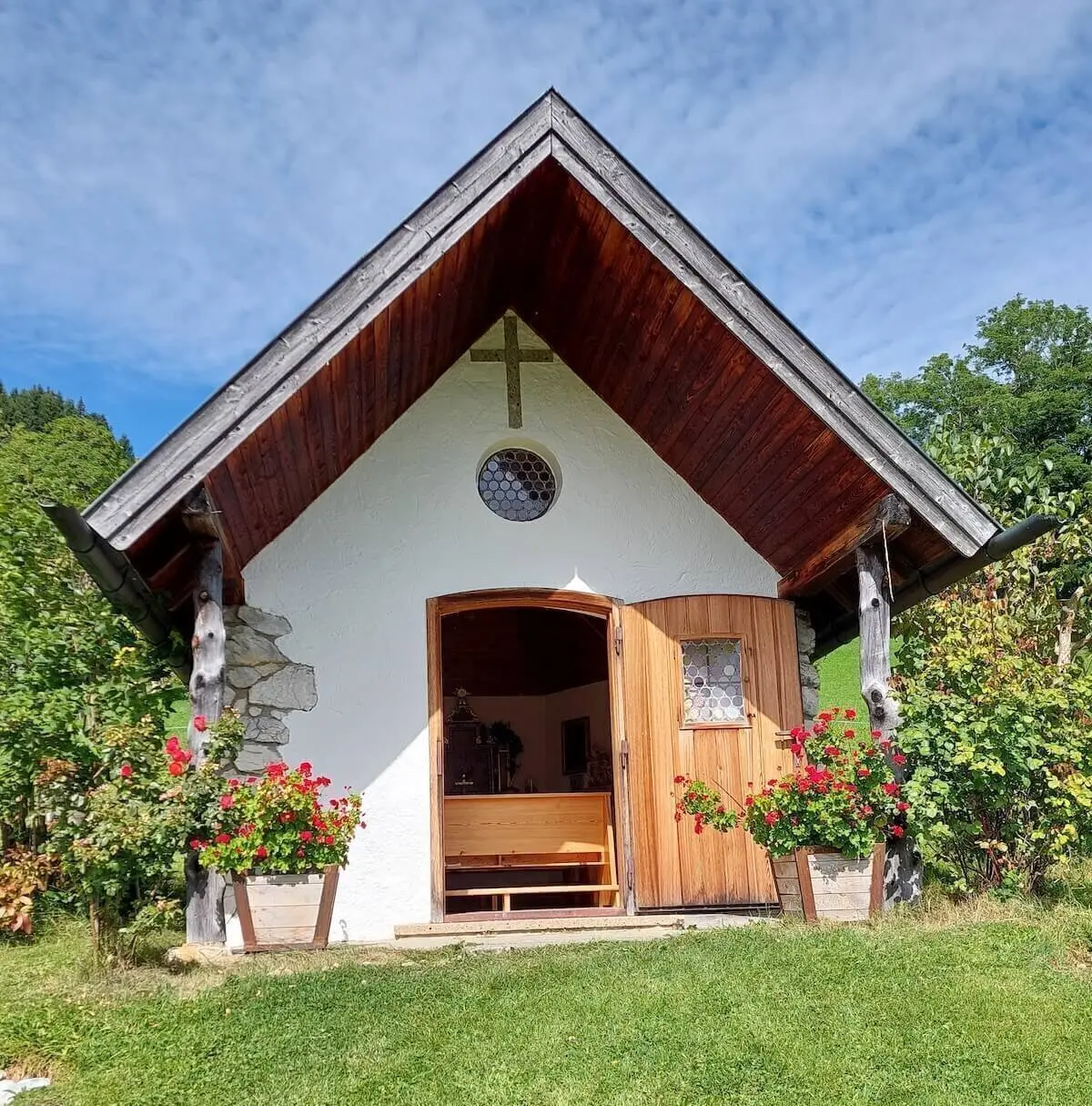 Weiße Kapelle mit dunklem Holzdach und roten Geranien, umgeben von Bäumen unter leicht bewölktem Himmel. | © DAV Markt Schwaben | Foto Hubert Inhofer