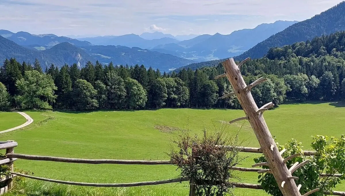 Schräge Holzstruktur lehnt am Zaun vor Wiese und Wald, im Hintergrund blaue Berge unter lockerem Wolkenhimmel. | © DAV Markt Schwaben | Foto Hubert Inhofer