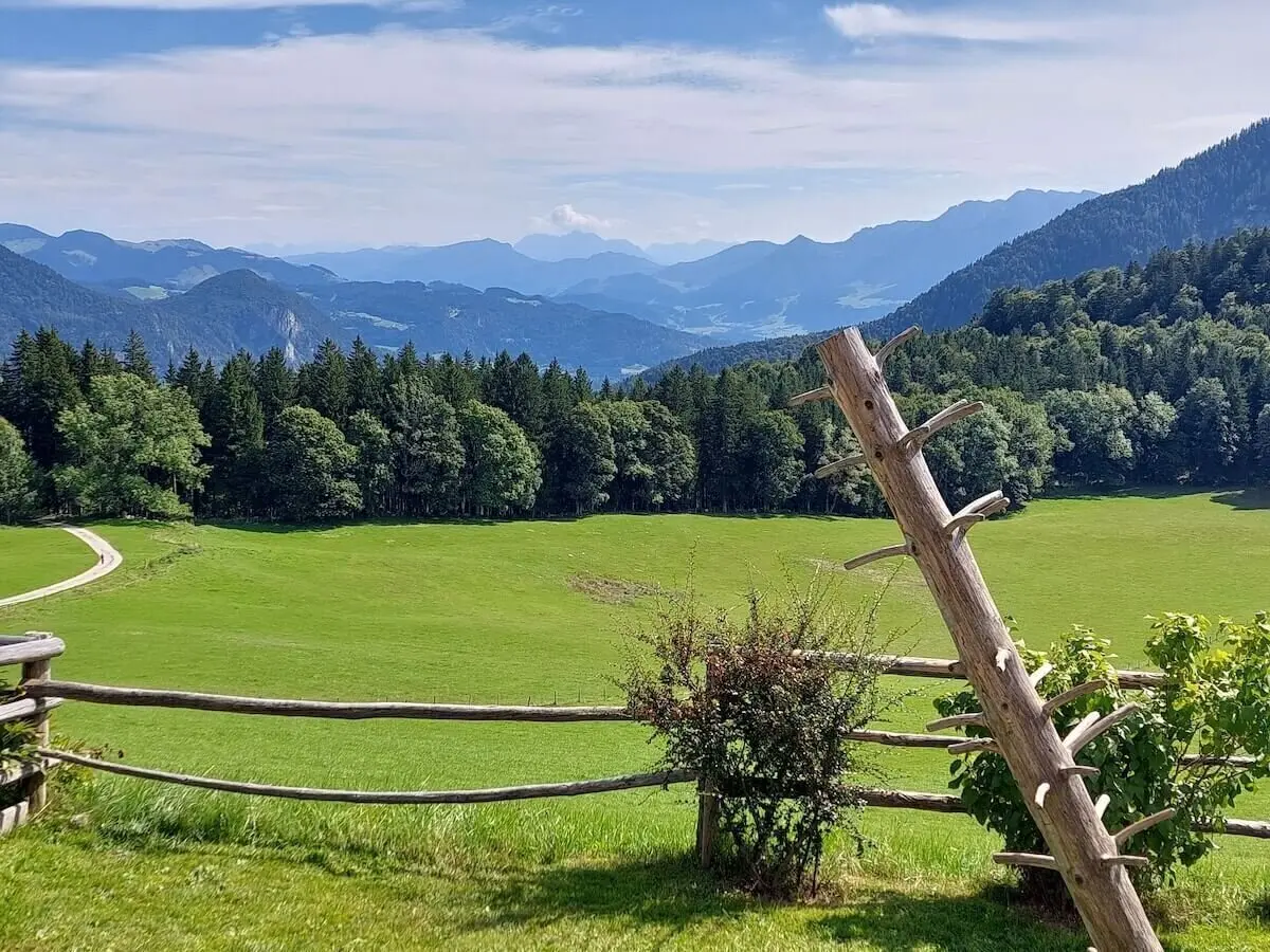 Schräge Holzstruktur lehnt am Zaun vor Wiese und Wald, im Hintergrund blaue Berge unter lockerem Wolkenhimmel. | © DAV Markt Schwaben | Foto Hubert Inhofer