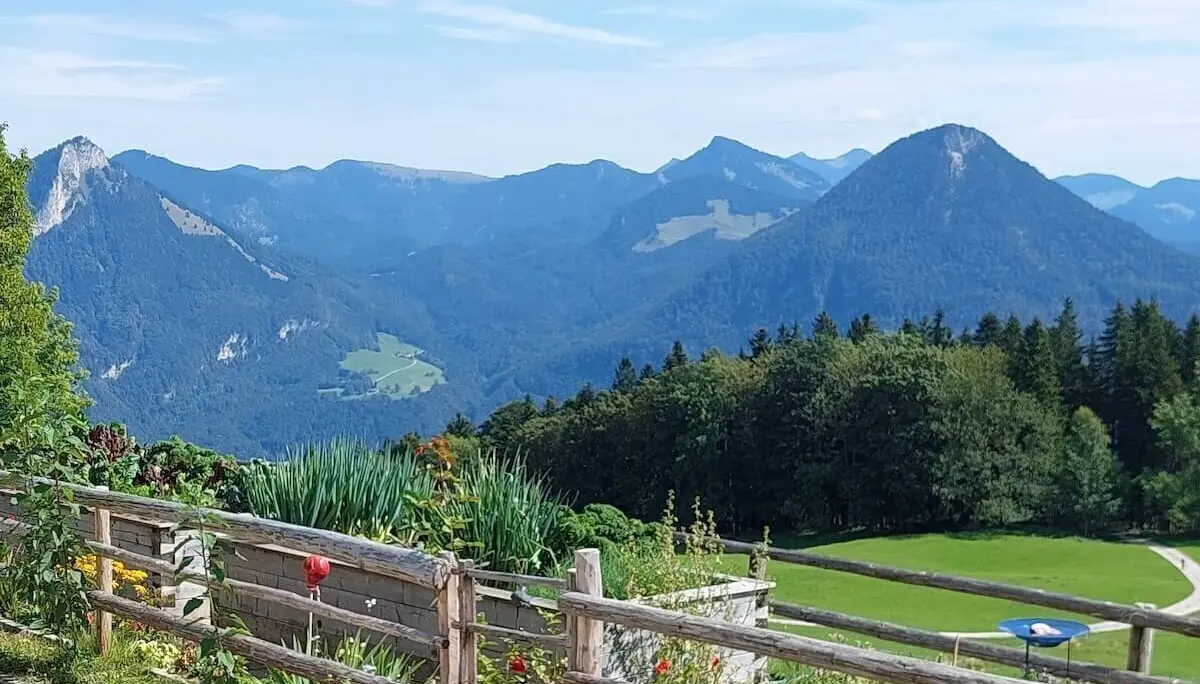 Blumenbeet und blauer Sonnenschirm vor Wiese und bewaldeten Bergen, eingerahmt von Holzzaun und Gartenweg. | © DAV Markt Schwaben | Foto Hubert Inhofer