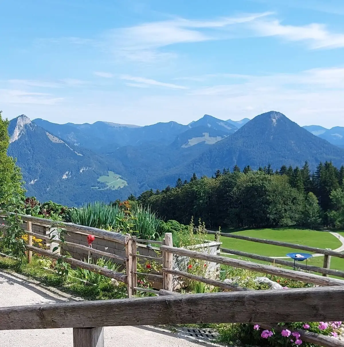 Blumenbeet und blauer Sonnenschirm vor Wiese und bewaldeten Bergen, eingerahmt von Holzzaun und Gartenweg. | © DAV Markt Schwaben | Foto Hubert Inhofer