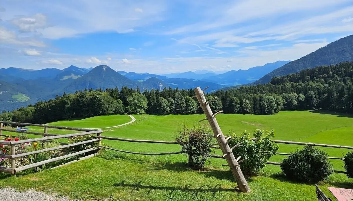 Holzzaun mit schräger Leiterstruktur vor Wiese mit Büschen, dahinter Waldhügel und Berge unter Sommerhimmel. | © DAV Markt Schwaben | Foto Hubert Inhofer