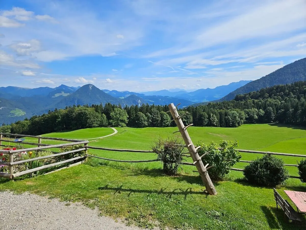Holzzaun mit schräger Leiterstruktur vor Wiese mit Büschen, dahinter Waldhügel und Berge unter Sommerhimmel. | © DAV Markt Schwaben | Foto Hubert Inhofer