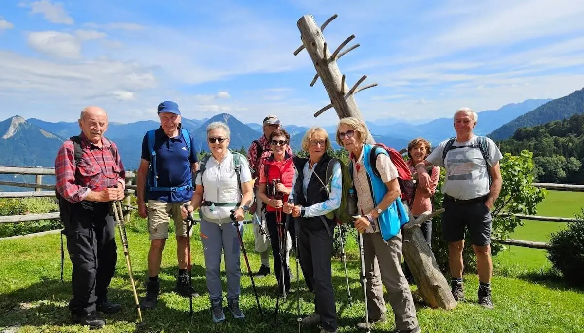 Neun Wandernde stehen auf Wiese vor Holzskulptur und Zaun, dahinter Hügel und Berglandschaft bei Sonnenschein. | © DAV Markt Schwaben | Foto Hubert Inhofer